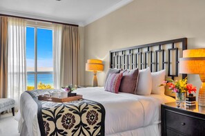Una habitación de hotel con cama, lámparas de noche, mesita de noche, ventana con cortinas y vista al océano.