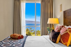 Hotellihuone, jossa on suuri ikkuna, kuviollinen vuodevaatteilla varustettu sänky, lamppu ja näkymä merelle.