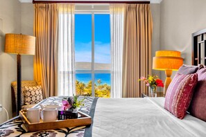A hotel room with a bed, two lamps, a tray with mugs, a vase with flowers, and a view of palm trees through the window.