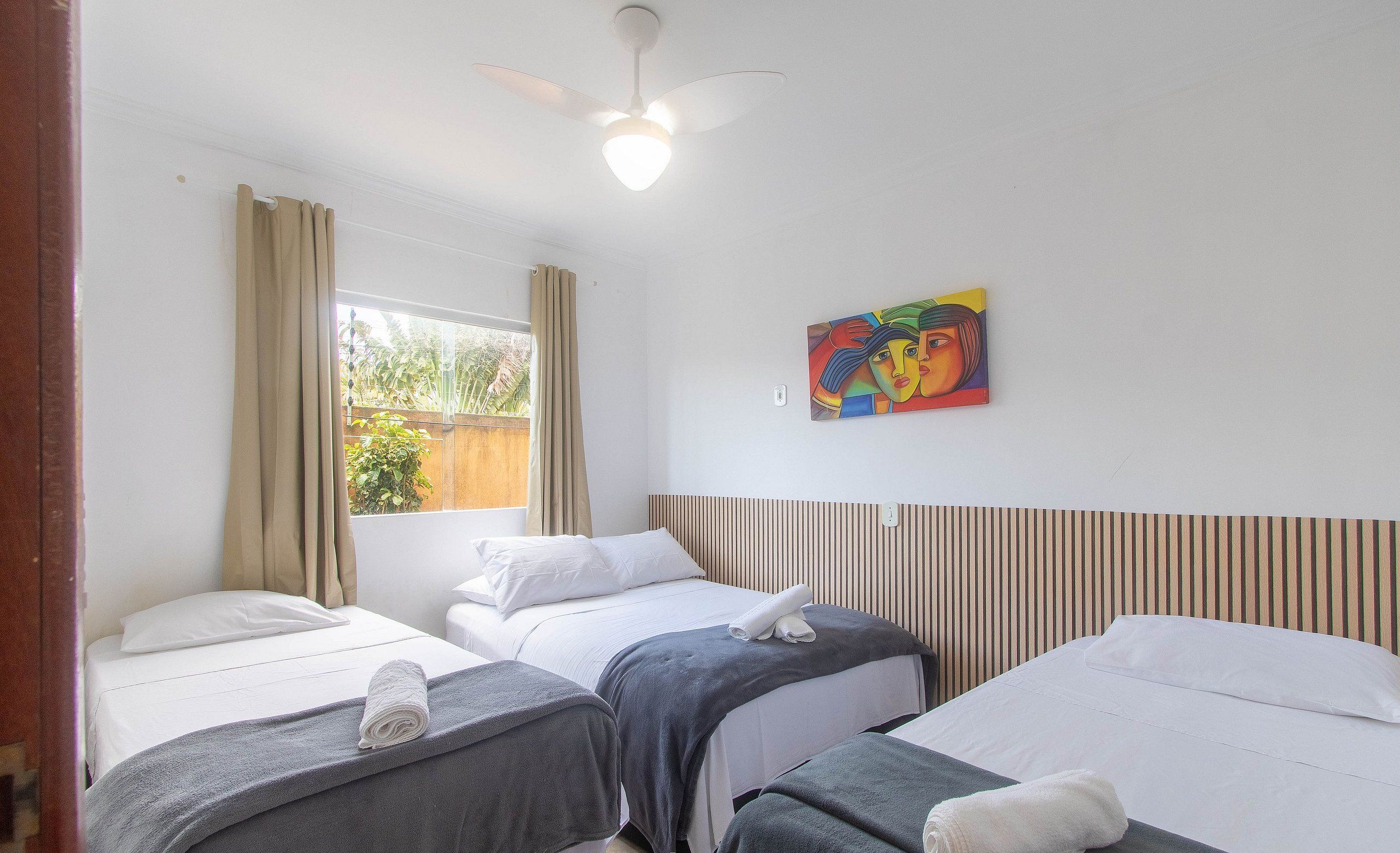Quarto de hotel com três camas, um ventilador de teto, uma janela com cortinas e um quadro na parede.