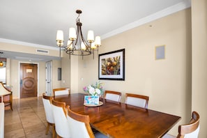 A dining room with a wooden table, white chairs, a chandelier, and a framed picture on the wall. - Honua Kai | Ocean View 8 BR, Sleeps 22 | Car Incl. w/ 6+ Nights | HKK ML-1457 by KBM (Lahaina)