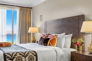 A hotel room with a large bed, two bedside lamps, a nightstand with a vase of flowers, and a view of the sea through a window with curtains. - Honua Kai | Ocean View 8 BR, Sleeps 22 | Car Incl. w/ 6+ Nights | HKK ML-1457 by KBM (Lahaina)