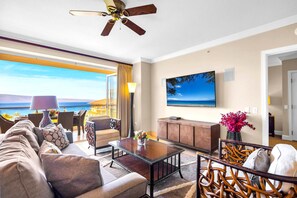 Amplia sala de estar con un gran ventanal con vistas a un paisaje marino, ventilador de techo, televisor de pantalla plana y una zona de estar confortable.