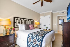 A hotel room with a bed, two bedside lamps, a nightstand, a dresser, a ceiling fan, and a door leading to another room. - Honua Kai | Ocean View 8 BR, Sleeps 22 | Car Incl. w/ 6+ Nights | HKK ML-1457 by KBM (Lahaina)