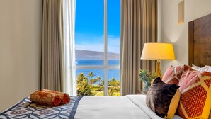 Ein Hotelzimmer mit großem Fenster, einem Bett mit gemusterten Betttextilien, einer Lampe und Blick aufs Meer.