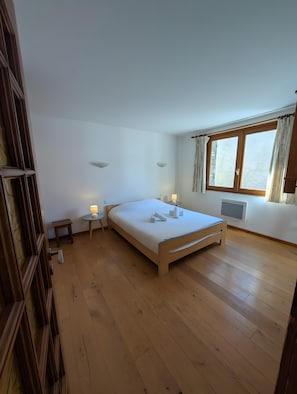 A bedroom with a wooden bed, a small bedside table with a lamp, a window with curtains, and a radiator.