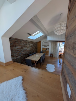 A modern loft-style room with a sloped ceiling, a dining area with a wooden table and chairs, a brick accent wall, and a large window with a skylight.
