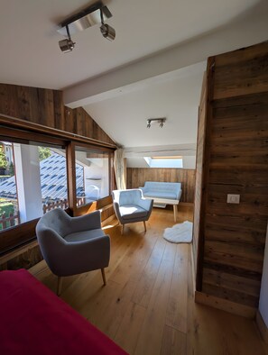 Un dormitorio tipo loft con techo inclinado, paredes de madera, un ventanal grande, una cama roja y un área de descanso con una silla y un sofá.