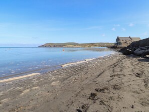 A beach with a rocky shoreline, a building with a steep roof, and calm water. - Ty Gwartheg (Newport)