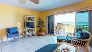 A living room with a TV, sofa, and chairs, a ceiling fan, and a balcony with a view. - Beachfront condo with washer dryer pool and hot tub dog friendly (Indian Rocks Beach)