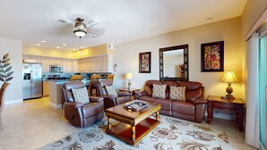 A living room with a brown leather sofa, a wooden coffee table, and a floral rug. There is a kitchen in the background with white cabinets and a microwave. - Welcoming Gulf front condo with pool and hot tub direct beach access (Indian Rocks Beach)