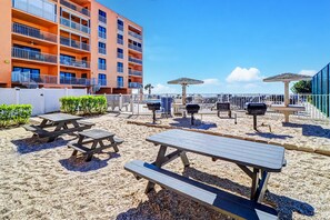 A beachside patio with picnic tables, barbecue grills, and umbrellas, adjacent to a multi-story building with balconies. - Bright and recently redone condo hot tub pool and tennis direct beach access (Indian Rocks Beach)