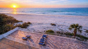 Blick auf einen Strand bei Sonnenuntergang, eine Terrasse mit Stühlen und eine Palme.