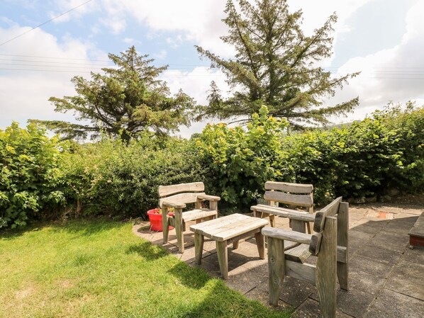 A patio area with wooden benches and a picnic table, surrounded by lush greenery and trees. - Ty Fferm (Newport)