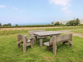 Una mesa de madera rústica y bancos ubicados en un campo cubierto de hierba, con una vista panorámica del mar y las colinas al fondo.