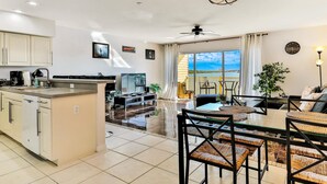 A modern kitchen with a central island, a dining area with a glass table and woven chairs, and a living room with a sofa and a large window offering a view of the outdoors.
