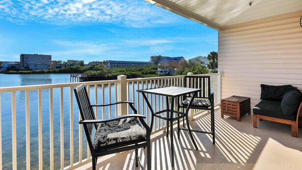 A balcony with a view of a lake, a table, two chairs, and a small wooden stool.