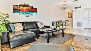 A modern living room with a sectional sofa, a coffee table, and a dining area with a table and chairs. There is a large painting on the wall and a plant in the corner.