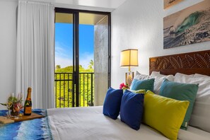 A hotel room with a bed, pillows, a nightstand with a lamp, a window with curtains, and a balcony view.