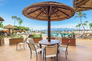 A poolside area with a round table and chairs, a thatched umbrella, and lounge chairs.