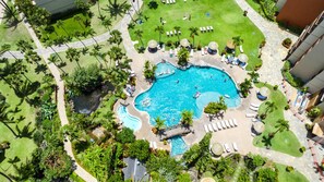 Vista aérea de un complejo turístico con piscina central, rodeado de palmeras, tumbonas y una pequeña cascada.