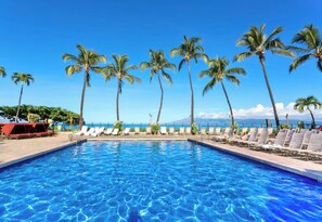 A swimming pool with clear blue water, surrounded by lounge chairs and palm trees, with a view of the ocean and a mountain in the background.