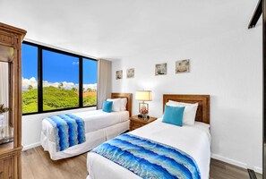 Quarto de hotel com duas camas, uma janela grande com vista para o verde, cabeceira de madeira e uma pequena mesa com abajur.