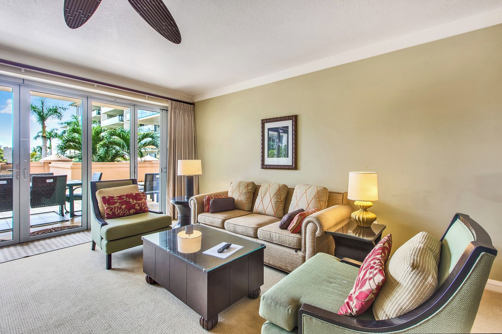 Habitación de hotel con sofá, sillón y una mesa de centro de vidrio. Hay una puerta corrediza de vidrio que da a un área exterior con vistas a palmeras.