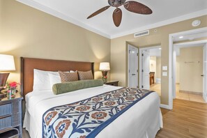 Habitación de hotel con una cama grande, un ventilador de techo, una mesita de noche con lámpara y una puerta que da a otra habitación.