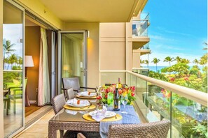 Un balcón con una mesa para dos personas, una silla y barandilla de vidrio con vistas a un paisaje tropical.