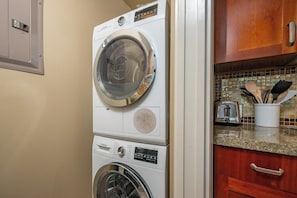 A stacked washer and dryer unit in a kitchen area with wooden cabinets and a tiled backsplash.