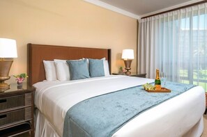 Ein Hotelzimmer mit einem großen Bett, zwei Nachttischen mit Lampen, einem Fenster mit Vorhängen und einer Champagnerflasche auf dem Bett.