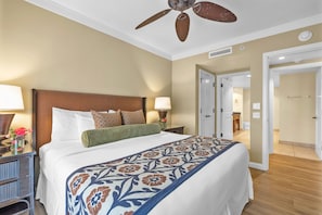 A hotel room with a large bed, a ceiling fan, a nightstand with a lamp, and a door leading to another room.
