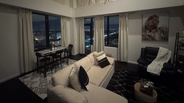 A high-ceiling living room with a sofa, a dining table, and a black leather chair. Large windows offer a city view.