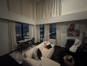 Un salón con techos altos, un sofá, una mesa de comedor y un sillón de cuero negro. Amplias ventanas ofrecen vistas a la ciudad.