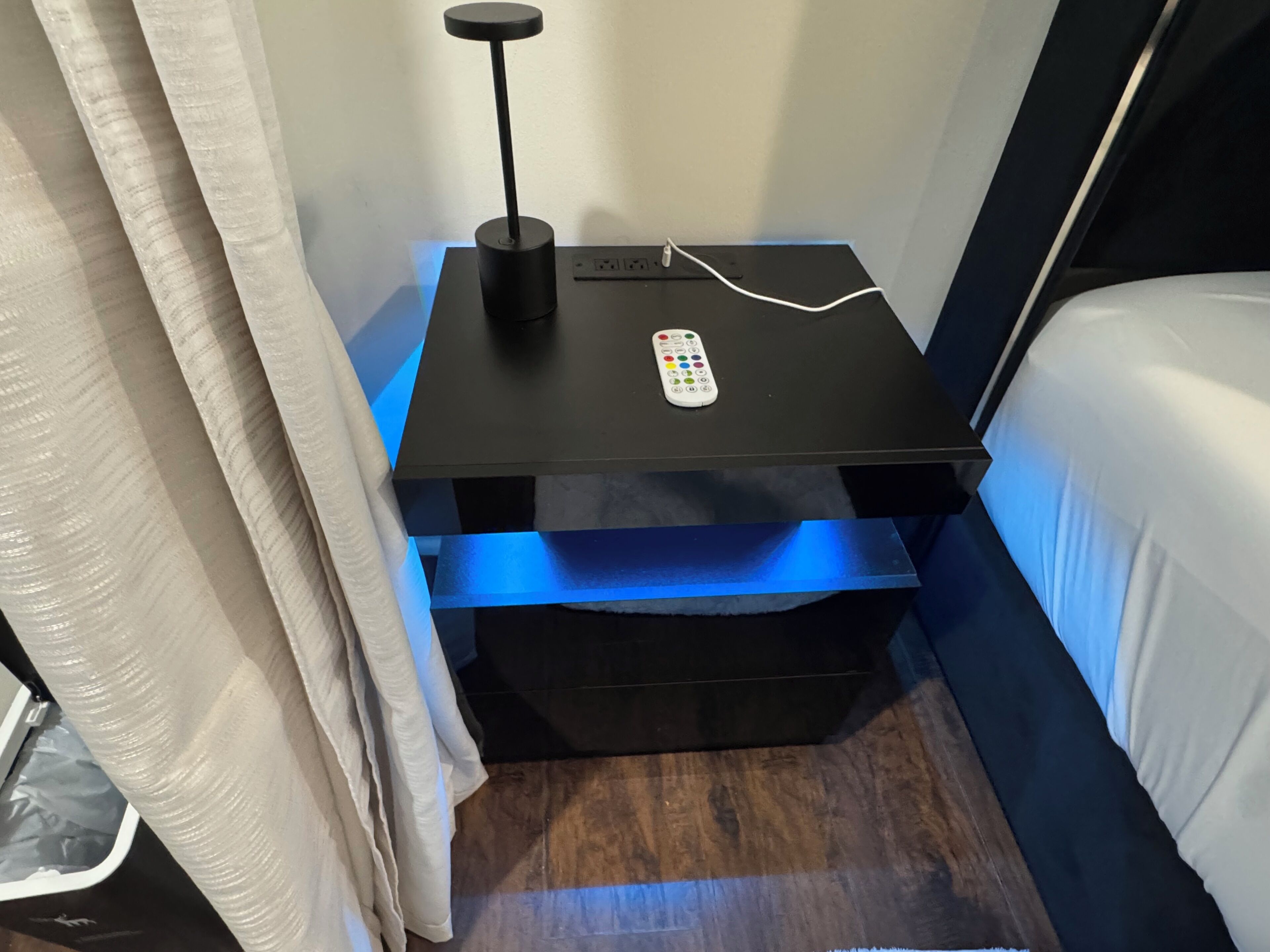 Una mesita de noche pequeña con un control remoto, un enchufe y un estante iluminado en azul debajo.