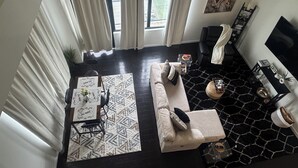 A modern living room with a sofa, a coffee table, and a TV. There are curtains, a rug, and a painting on the wall.