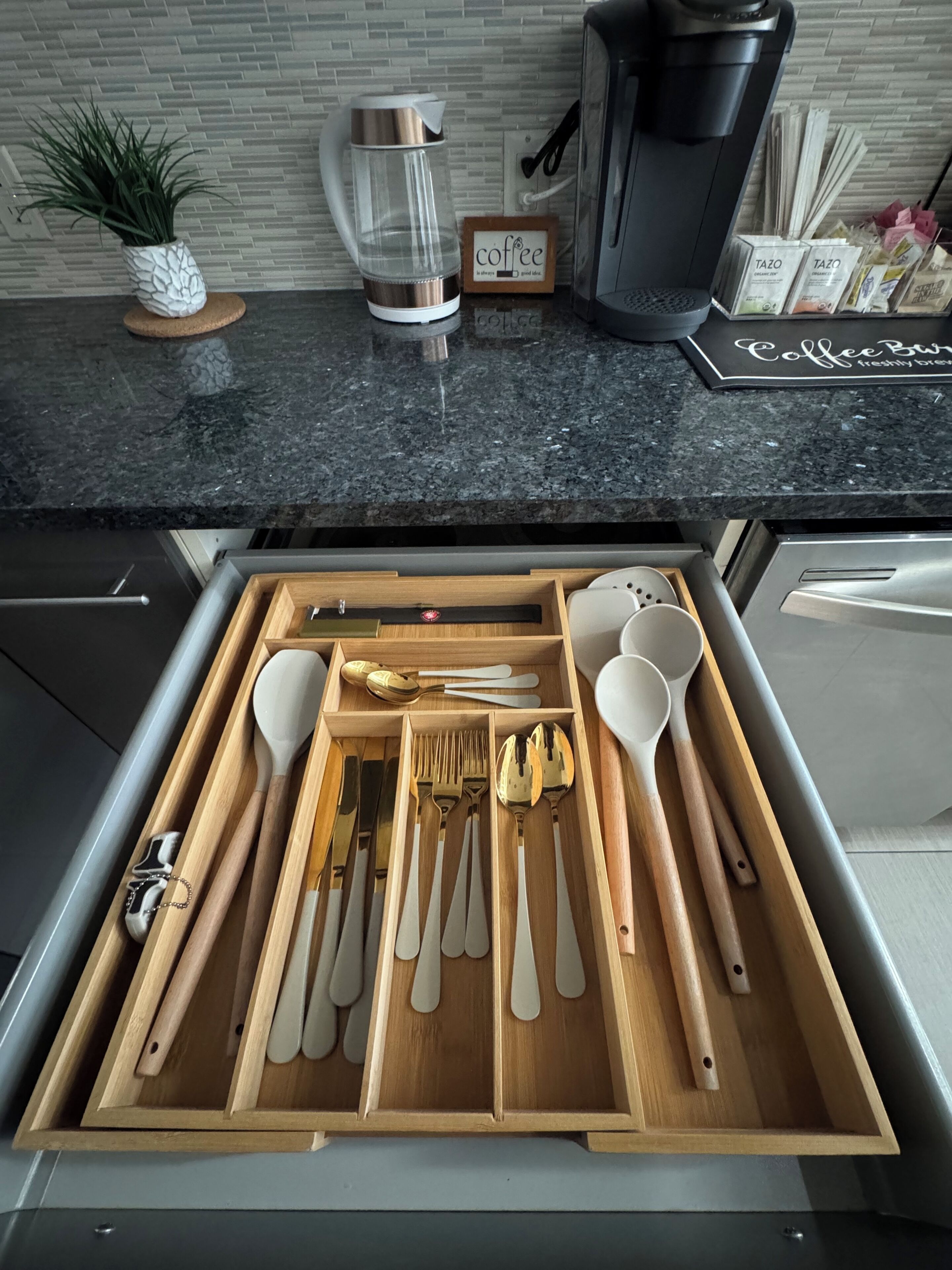 A kitchen with a coffee maker, a plant, and a drawer with utensils.
