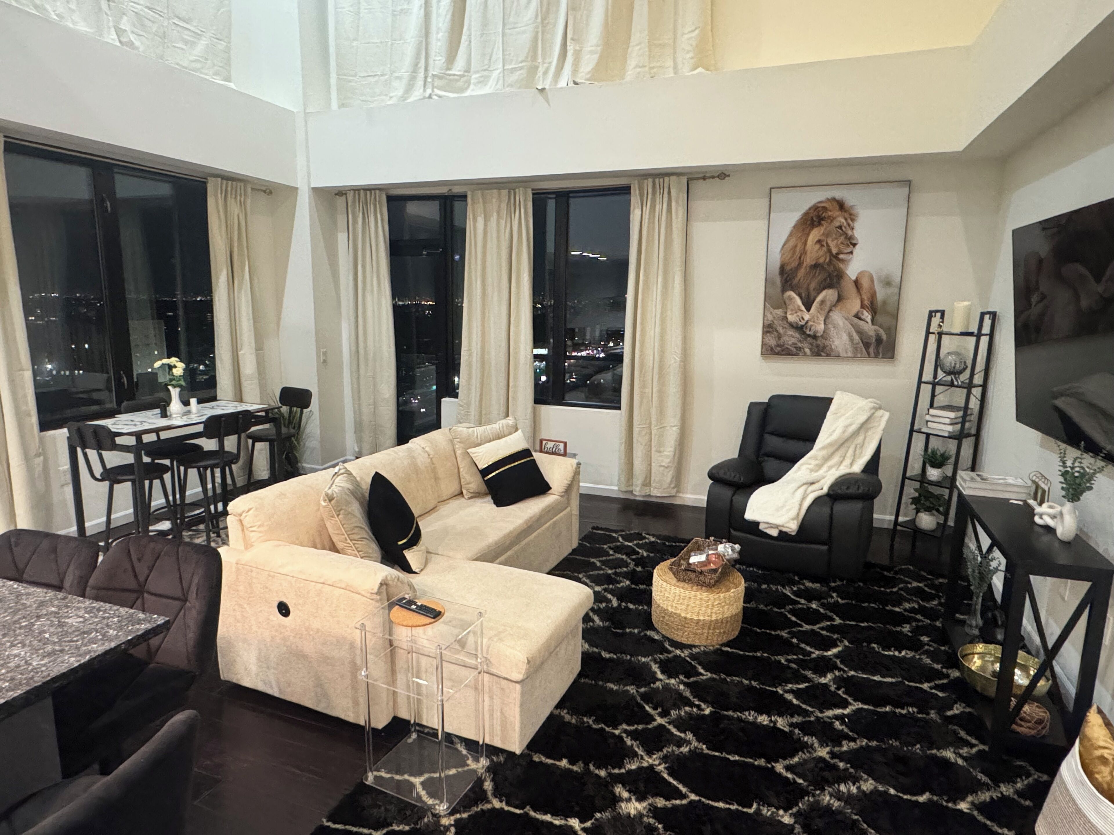Un salon moderne comprenant un canapé, un fauteuil noir, un coin repas avec une table et des chaises, et une grande fenêtre donnant sur la ville.