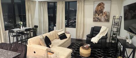Un salon moderne comprenant un canapé, un fauteuil noir, un coin repas avec une table et des chaises, et une grande fenêtre donnant sur la ville.
