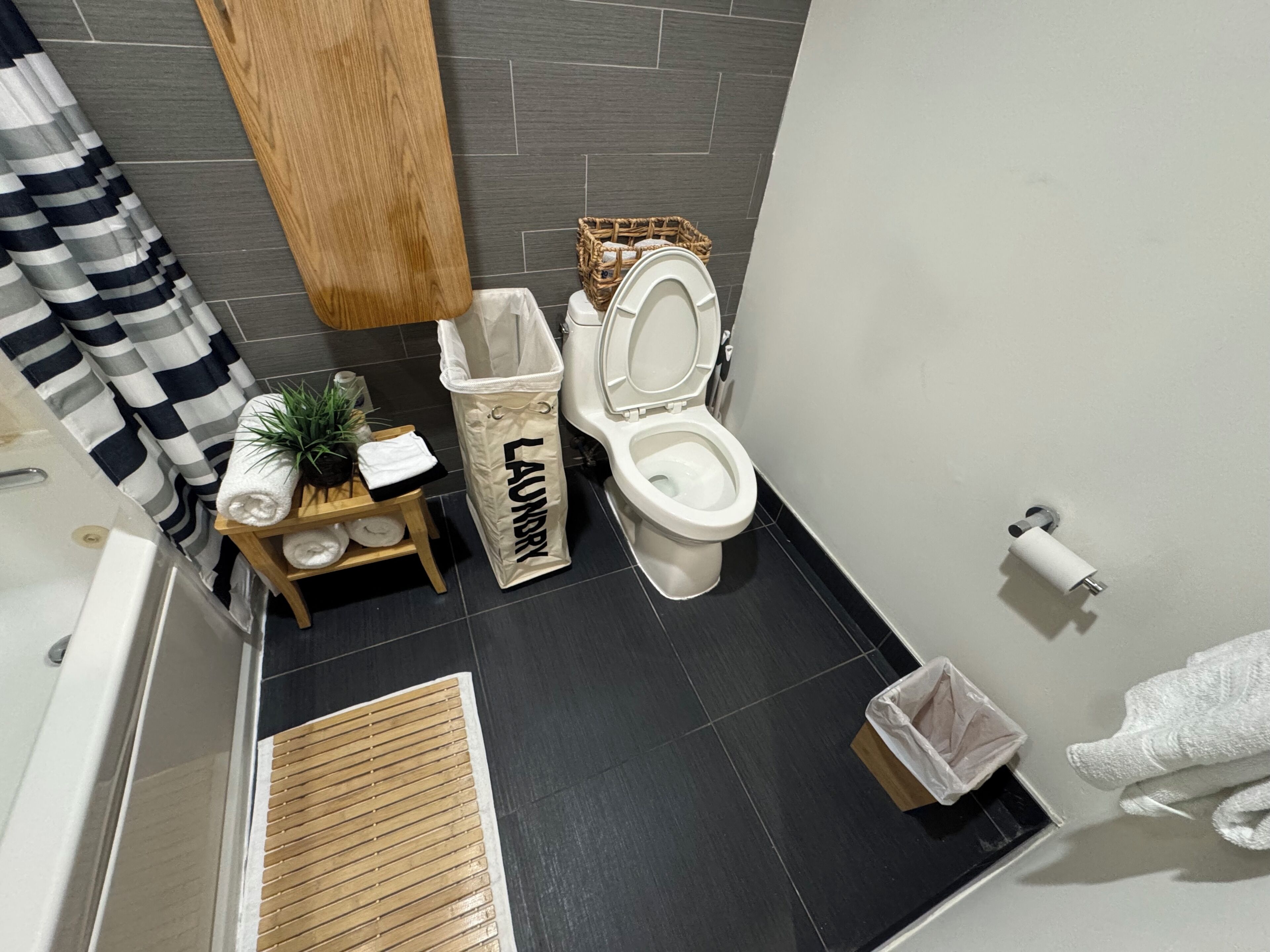 A bathroom with a toilet, a wooden shelf with towels, a laundry bag, and a small plant.