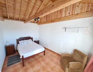 A bedroom with a wooden ceiling, a bed with a white bedspread, a nightstand with a lamp, a small dresser, and a checkered sofa.