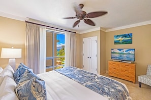 A hotel room with a bed, a ceiling fan, a TV, a wooden dresser, a chair, and a large window with curtains.