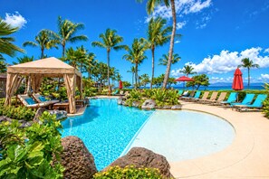 A pool area with lounge chairs and a canopy, surrounded by palm trees and a clear sky.
