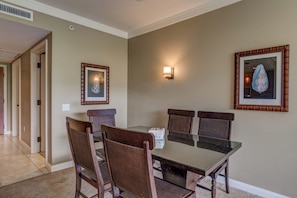 A dining area with a glass-top table, four chairs, two wall-mounted lamps, and framed artwork. - 2 Honua Kai Villas | Ocean Front | Sleeps 12 | Free Car w/ 6+ Nights | ML-1790 (Lahaina)
