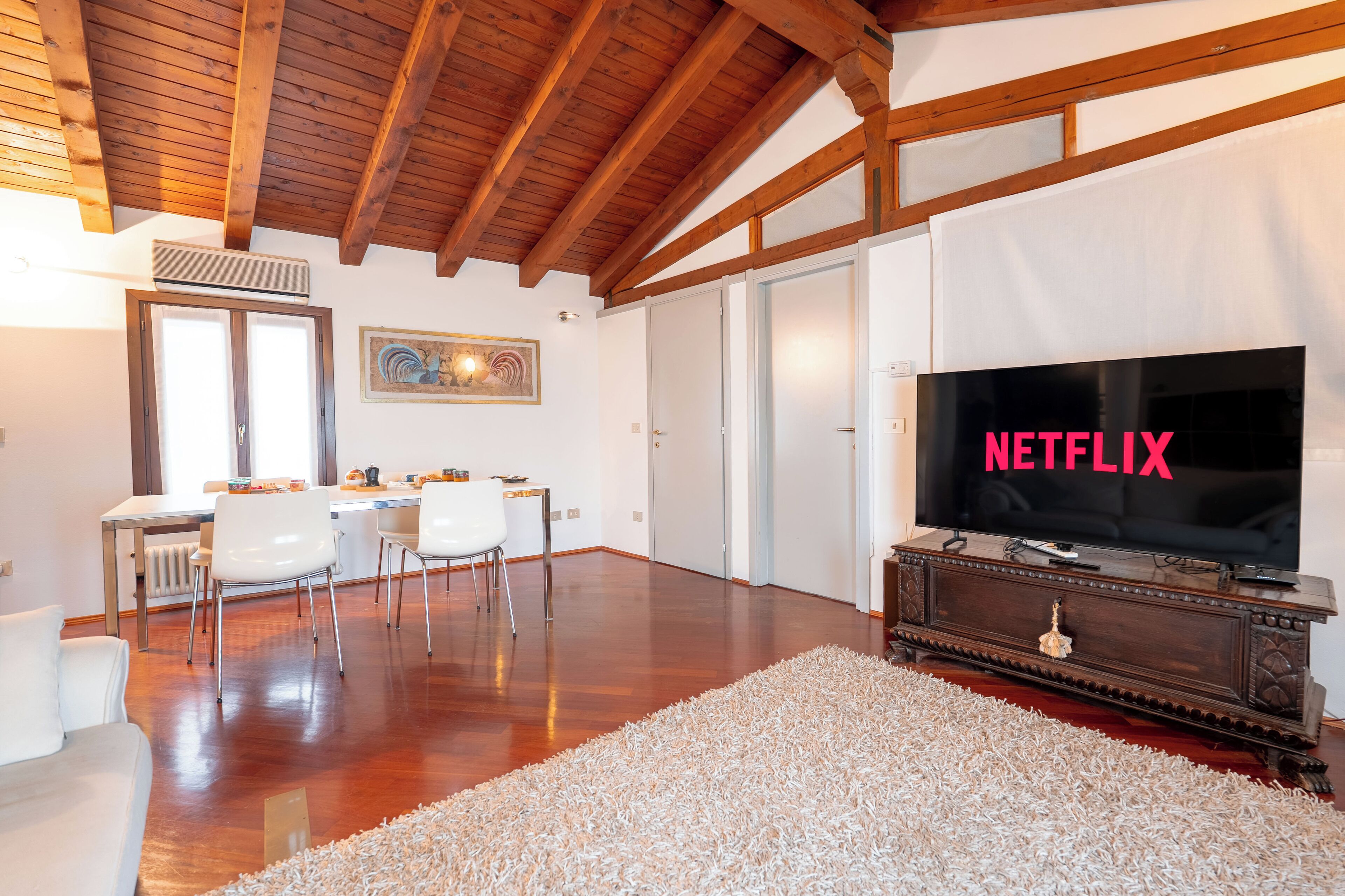 Ein großzügiges Wohnzimmer mit Holzbalkendecke, einem großen Flachbildfernseher, einer Essecke mit Stühlen und einem weißen Sofa.