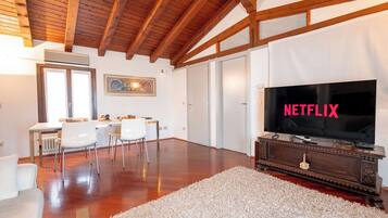 Ein großzügiges Wohnzimmer mit Holzbalkendecke, einem großen Flachbildfernseher, einer Essecke mit Stühlen und einem weißen Sofa.