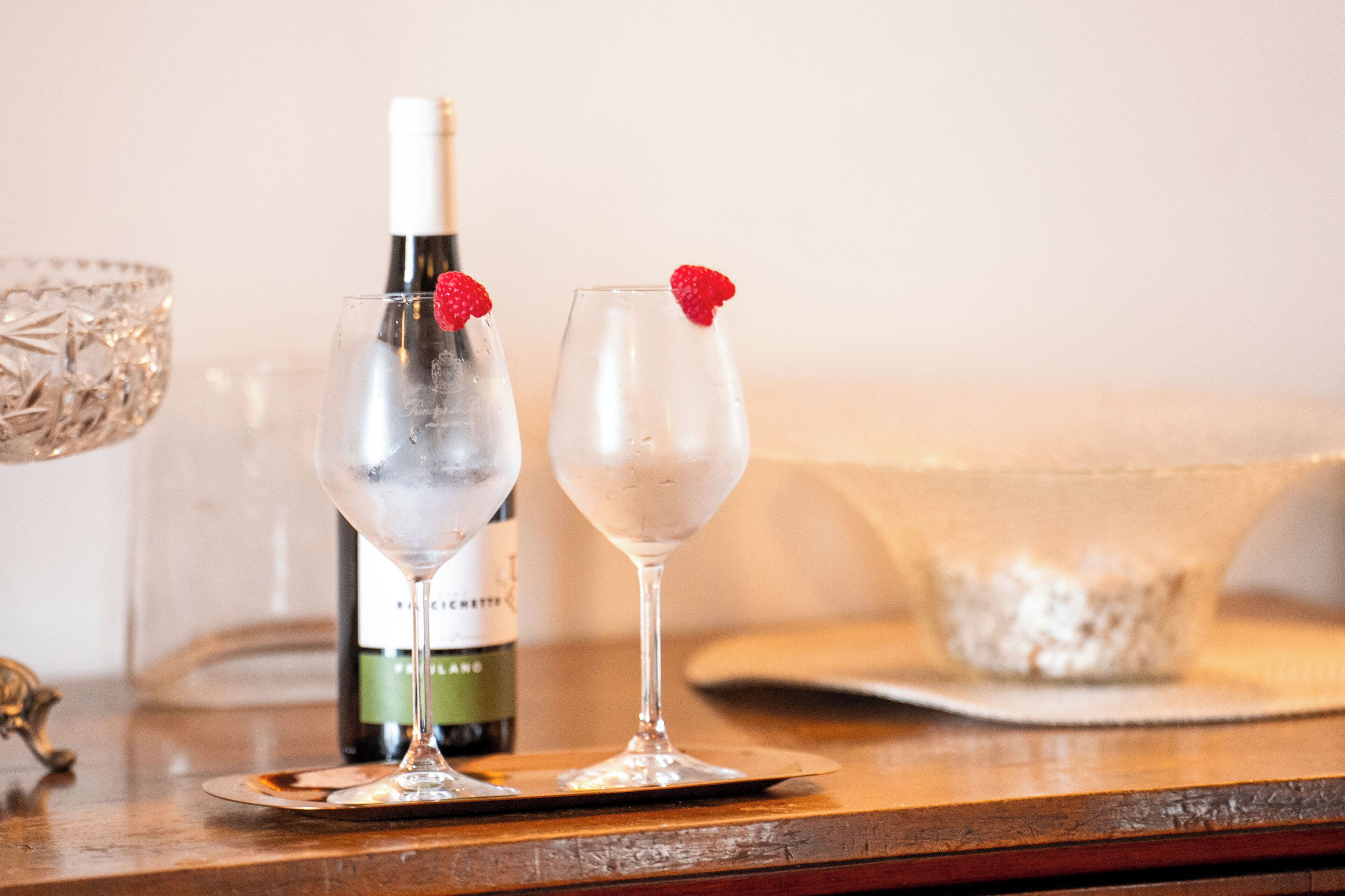 Two glasses of a clear beverage with a bottle of wine and a decorative bowl on a wooden table.