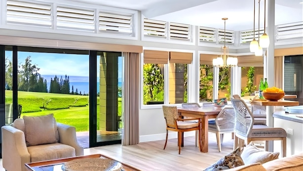 A spacious living area with a view of a golf course, a dining area with a table and chairs, and a kitchen area with a bowl of fruit on the counter. - NEW! KBM Resorts: Kapalua Ridge Villas KRV-2314 Sweeping Ocean, Golf/Mountain Views, w/Rental Car (Lahaina)