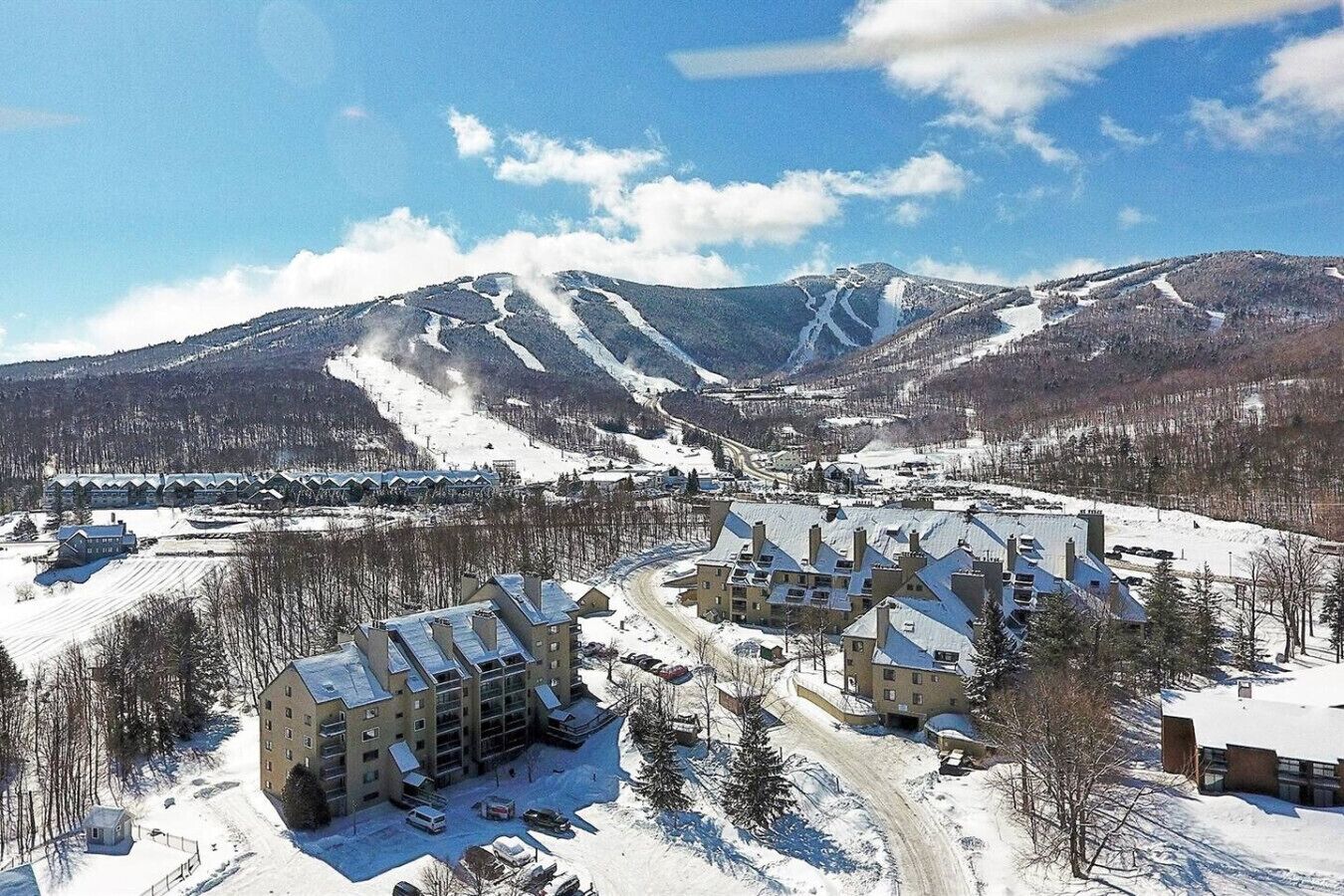 A snow-covered resort with multiple buildings, a ski slope, and mountains in the background.